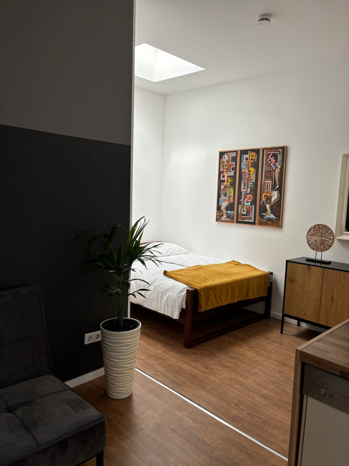 A well-lit bedroom features a spacious bed with crisp white linens, complemented by a mustard-colored throw. Adjacent to the bed, two framed artworks hang on the wall, above a wooden cabinet. A potted plant adds a touch of greenery to the room's neutral palette.