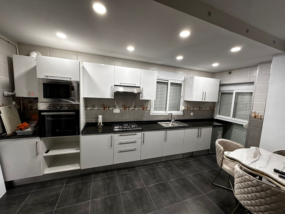 A modern kitchen equipped with sleek white cabinetry and a dark countertop is shown. Appliances include an oven, microwave, and gas stove. A dining area with a round table and stylish chairs is visible. Natural light enters through window panes partially covered by blinds.