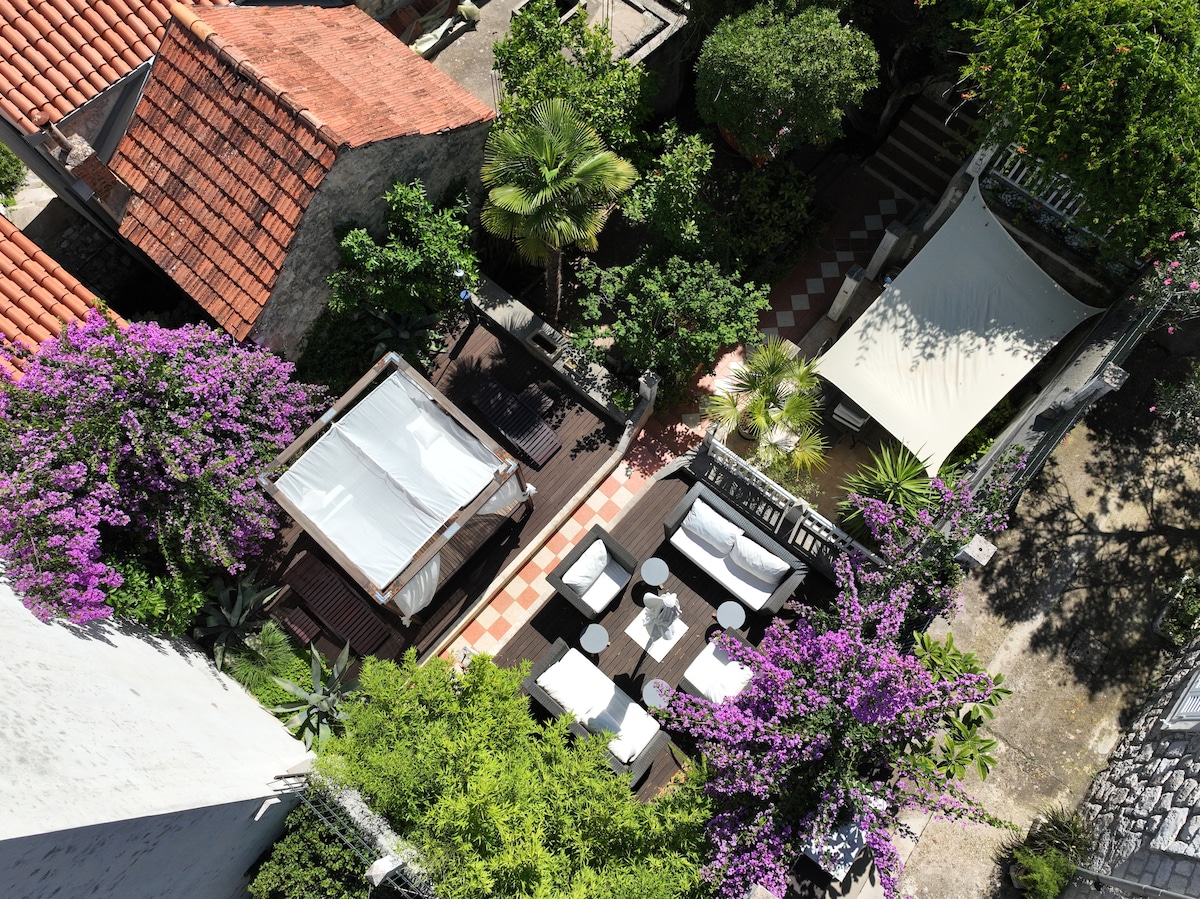 An aerial view captures a serene outdoor space adorned with vibrant purple flowers. Comfortable lounge chairs and a shaded seating area create inviting spots for relaxation, complemented by lush greenery and a sun canopy. The arrangement fosters a peaceful atmosphere, ideal for outdoor gatherings.