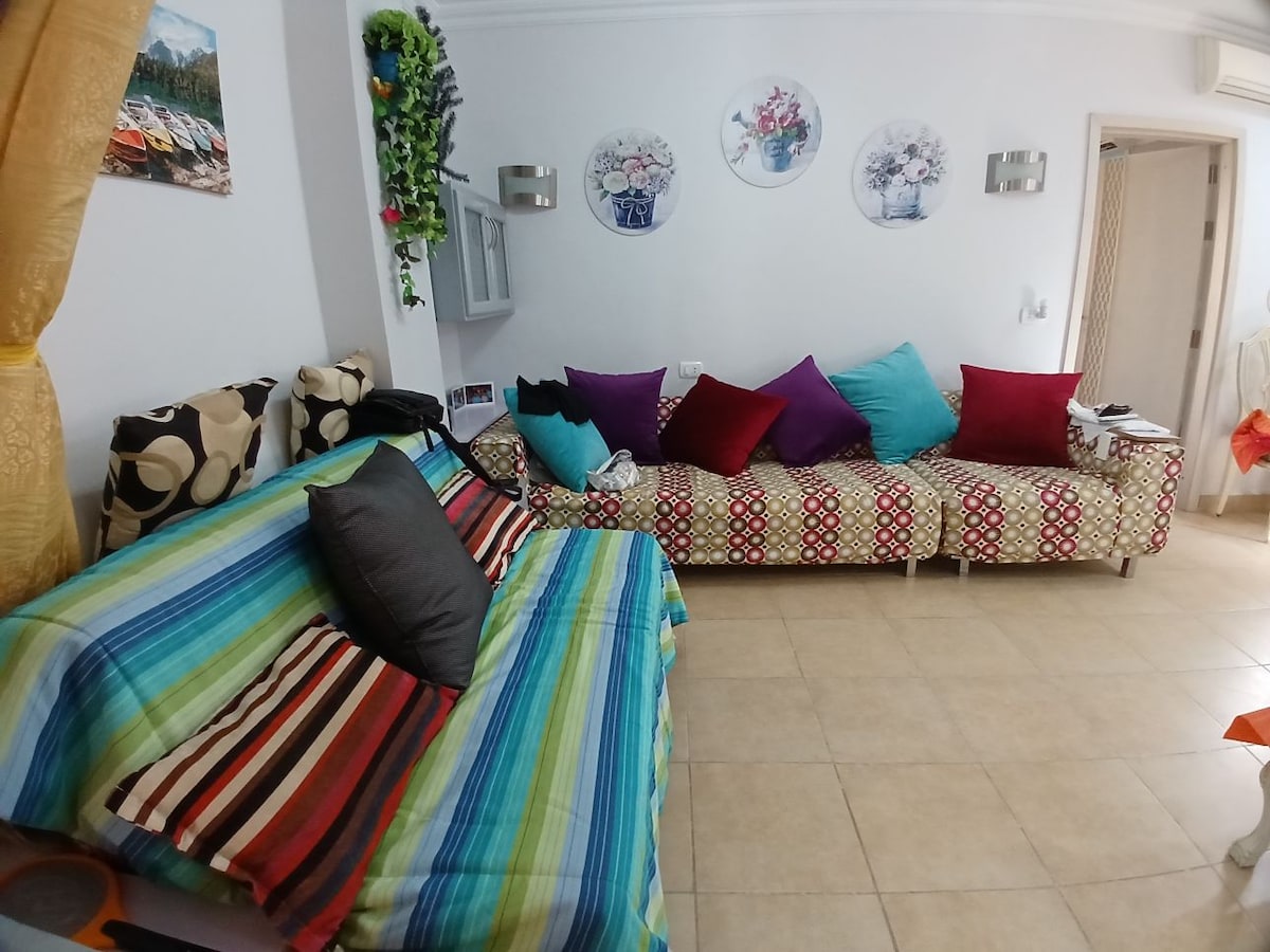 A welcoming living area features a patterned sofa adorned with colorful cushions, complemented by a striped blanket draped over a second, smaller sofa. Wall decor includes circular art pieces and greenery, while tile flooring enhances the bright and cheerful atmosphere.