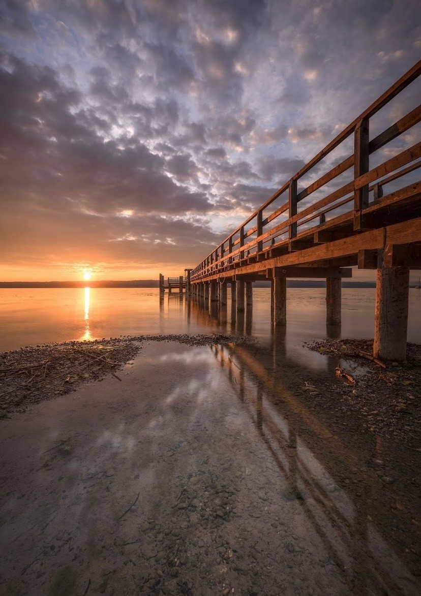 A wooden pier extends over calm waters, reflecting the soft hues of a sunset and cloudy sky. The sun is positioned low on the horizon, casting a warm glow, while the surrounding tranquility is highlighted by the stillness of the water.