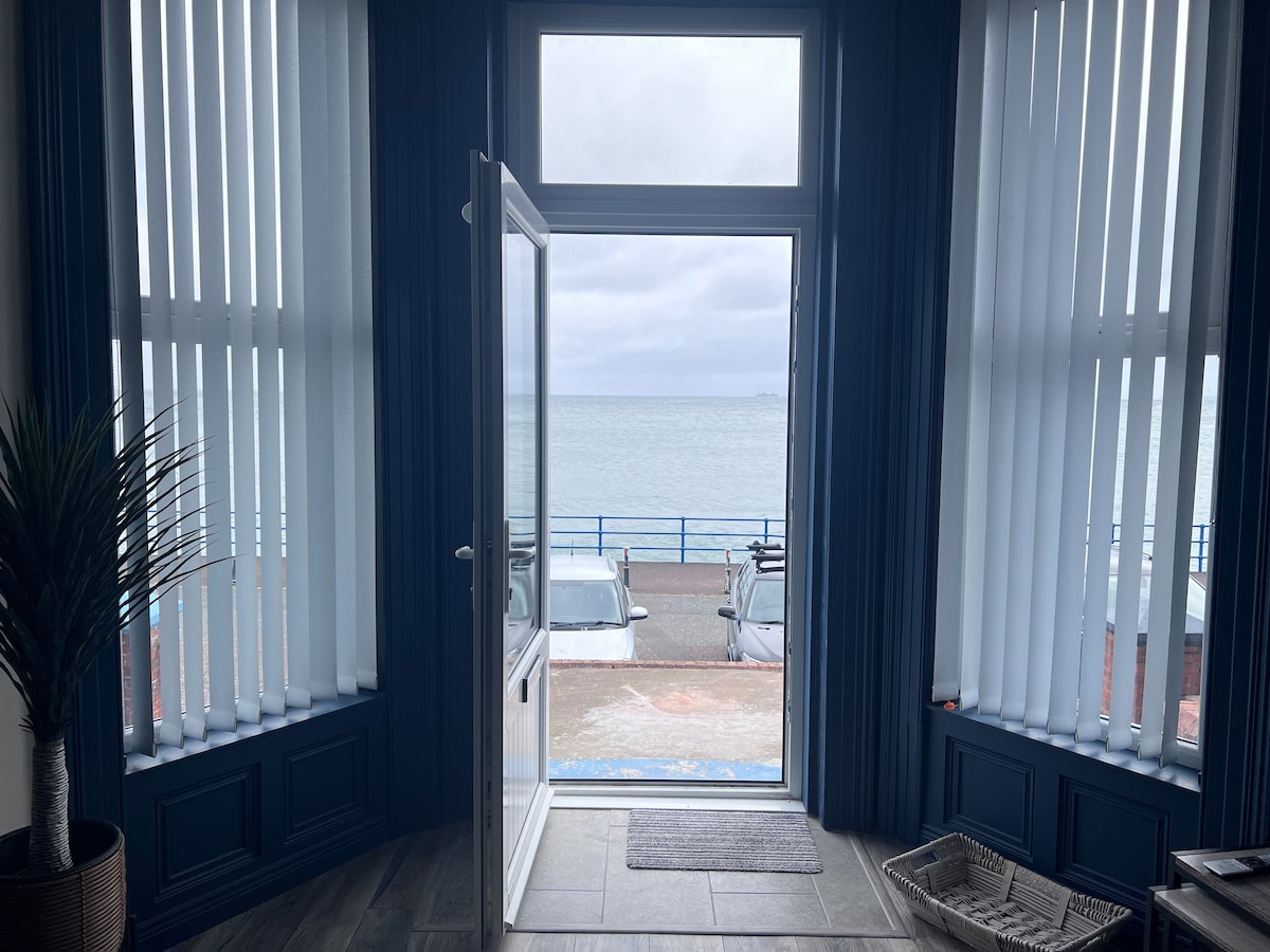 A door stands open, revealing a view of the sea beyond. Large windows with vertical blinds frame the scene, allowing soft, diffused light to enter. The floor features dark tiles, and a welcome mat is positioned at the entrance.