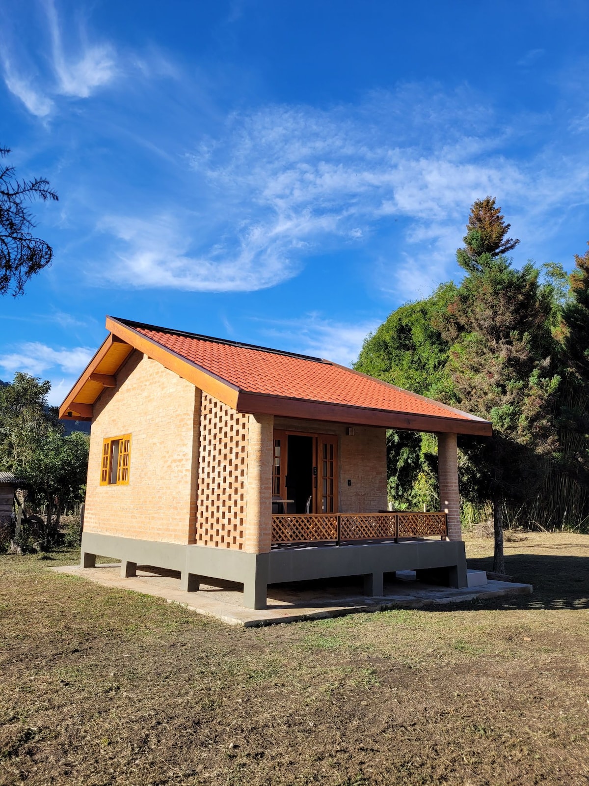 A cozy, newly constructed chalet is showcased, featuring rustic brick walls and a slanted red roof. A small front porch with lattice details offers a welcoming entryway, surrounded by a grassy area and trees in the background, providing a sense of tranquility.