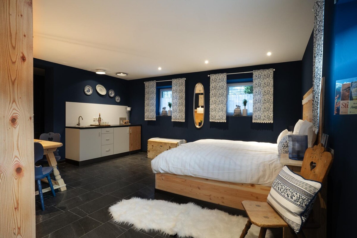 A spacious room features a large wooden bed with white linens. Soft rugs are placed on a dark stone floor. A dining area with a round table and four blue chairs sits in one corner. Natural light enters through two windows adorned with patterned curtains.