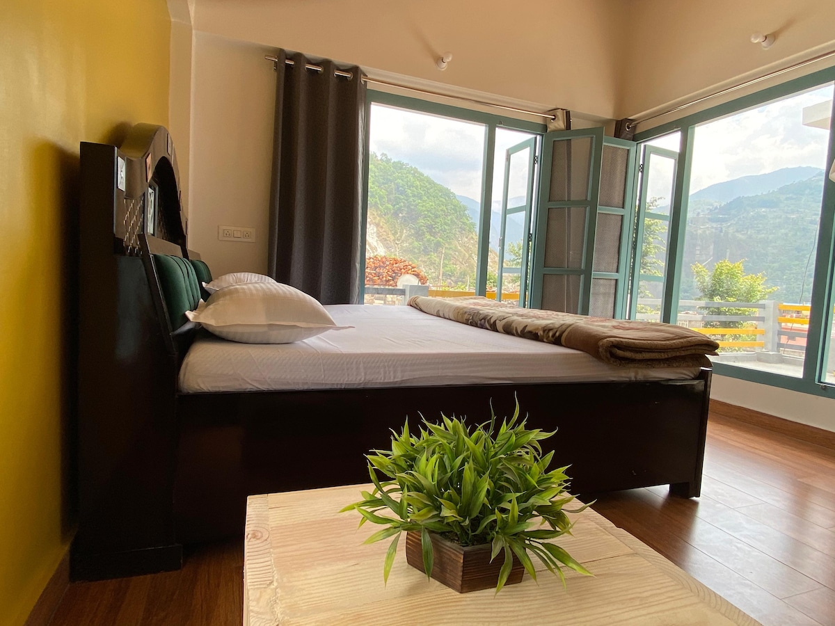 A sleeping area features a spacious bed adorned with soft linens and a folded blanket. Large windows invite natural light and offer views of the surrounding mountains. A small green plant rests on a wooden table, contributing to the serene ambiance.