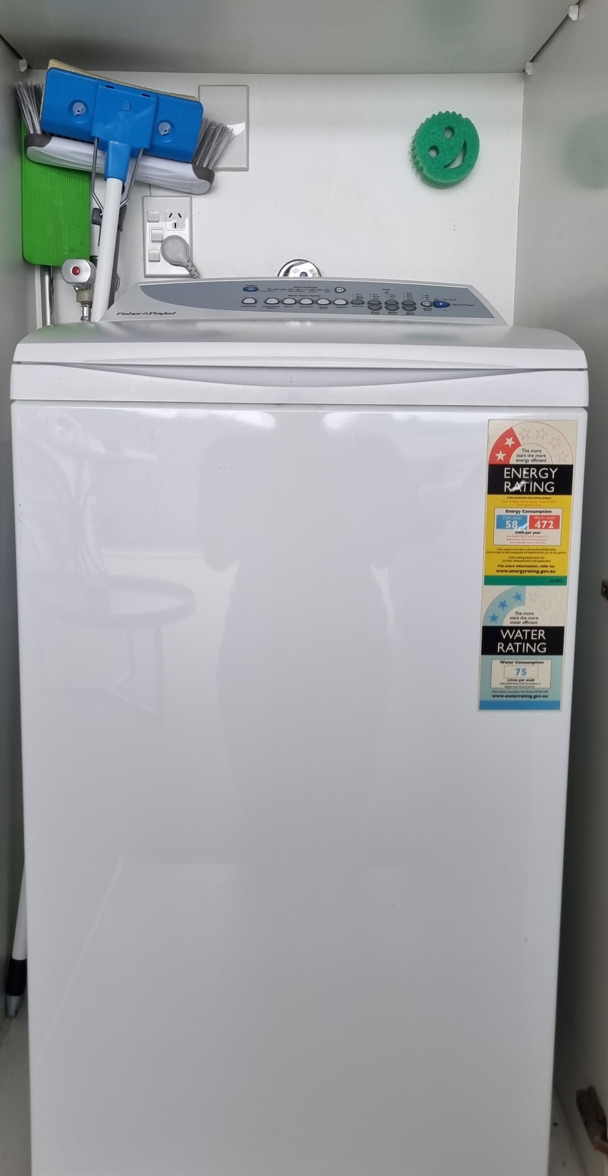 A top-loading washing machine is positioned in a compact utility space. Labels indicate energy and water efficiency ratings. Cleaning supplies, including brooms and a mop, are stored beside the machine, contributing to a tidy laundry area.