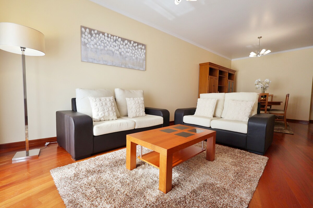 A spacious living area features two light-colored sofas against a neutral wall, complemented by a wooden coffee table at the center. A soft rug covers the floor, while a floor lamp adds warm light. In the background, a dining area with a wooden table and chairs is visible.