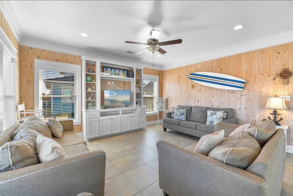 The living area is characterized by warm wood paneling and neutral-toned furnishings. A ceiling fan circulates air above two sofas and an armchair. A wall unit holds a large television, while decorative elements include a surfboard positioned above. Natural light filters in through the windows.