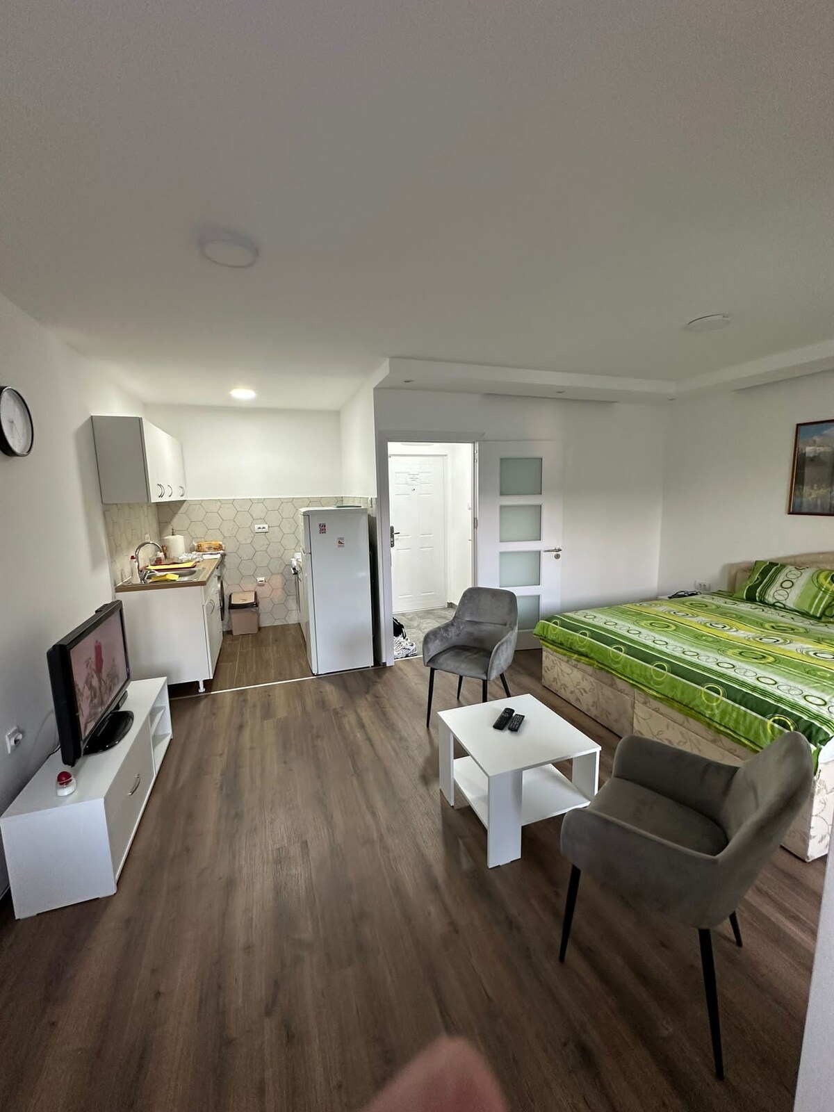 A bright and open living space features a spacious bed adorned with green bedding near a small dining area. A compact kitchen is present, along with a television and a coffee table positioned in front of two upholstered chairs. Natural light fills the room.