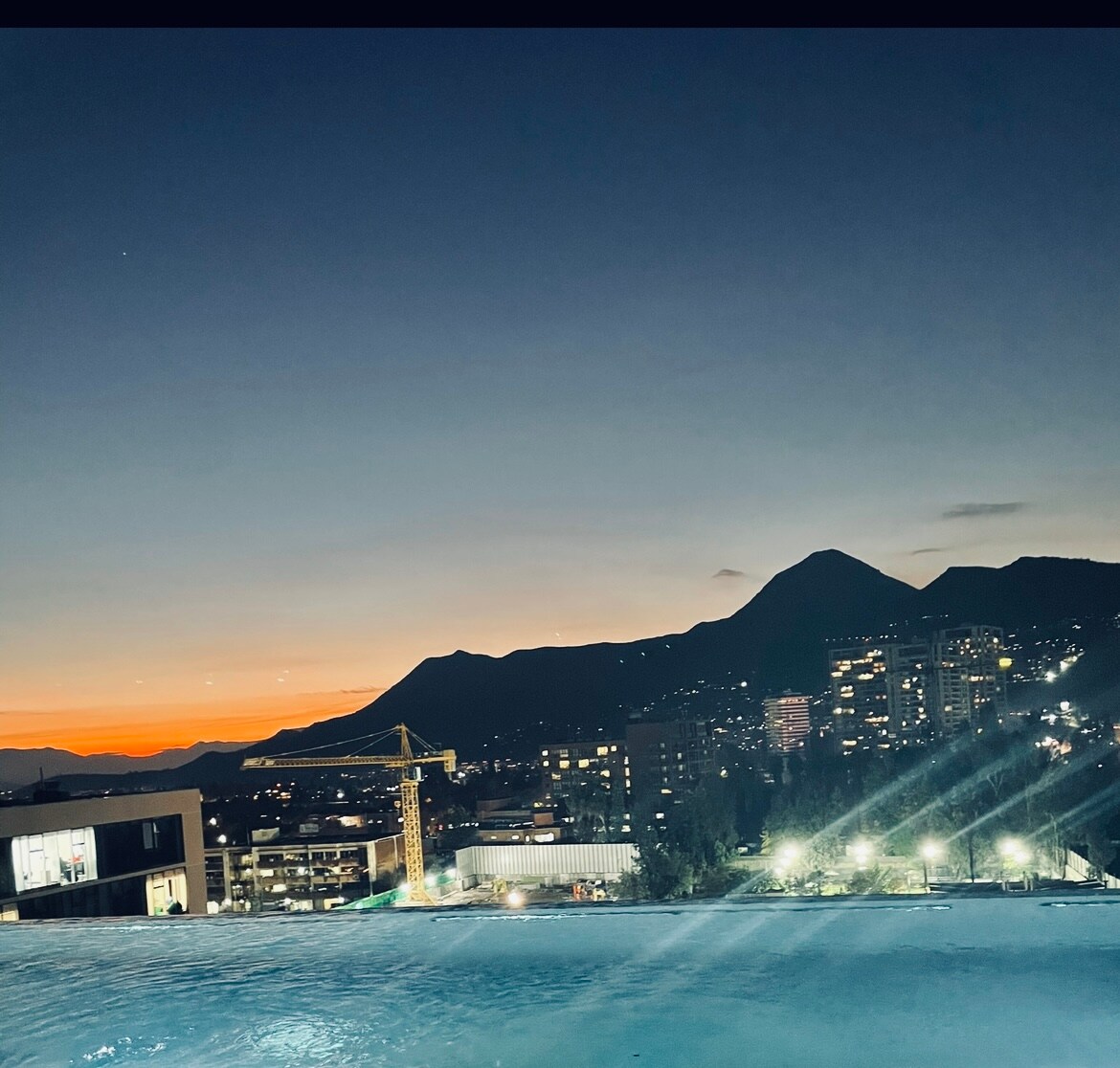 A panoramic view captures the evening sky as it transitions from orange to deep blue. Silhouetted mountains stand in the background, framed by softly glowing city lights. The tranquil water's edge reflects the colors of the sunset, enhancing the serene atmosphere.