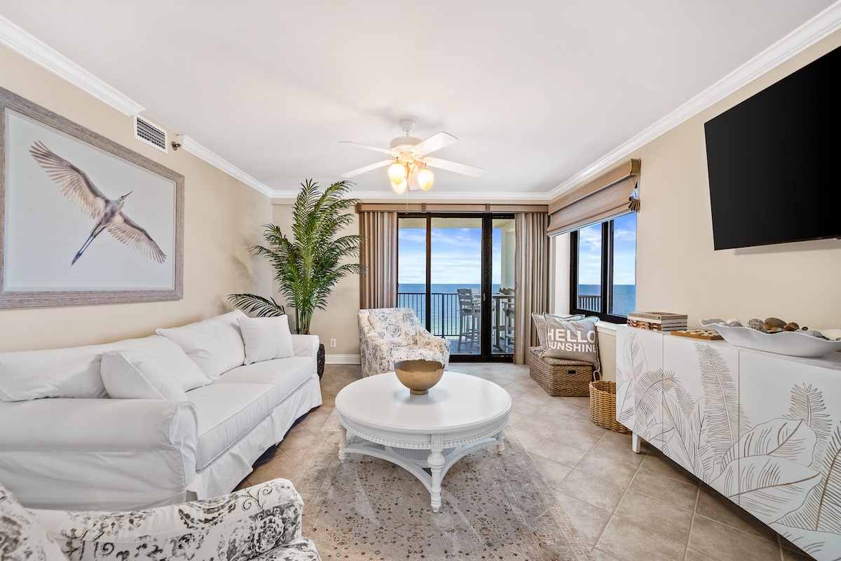 A light-filled living area features a comfortable white sofa and a decorative armchair, arranged around a round coffee table. Large glass doors offer views of the beach, while a decorative bowl sits atop the table. A ceiling fan and tropical plant add a relaxed touch.