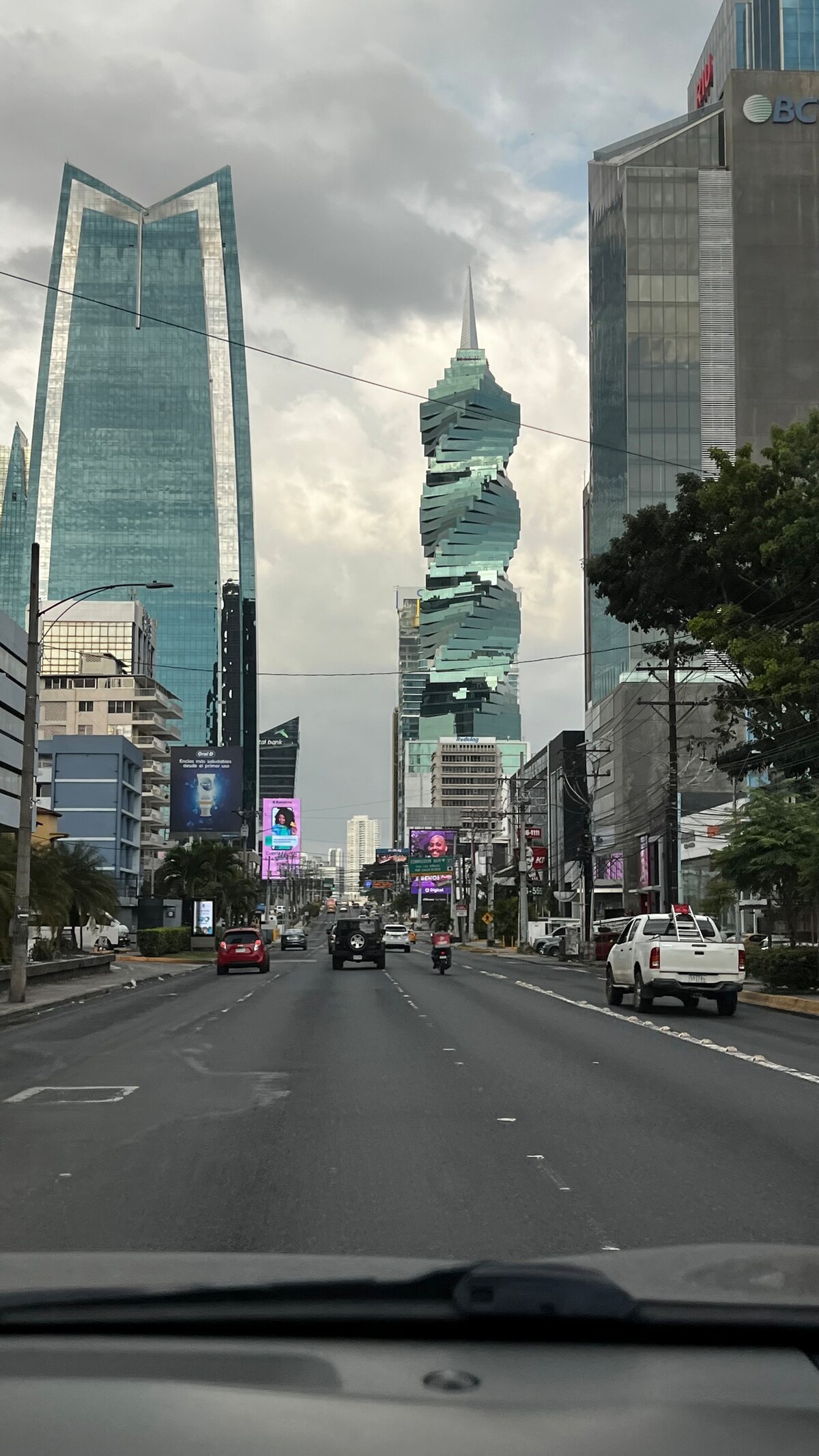 A city street is captured, showcasing modern skyscrapers, including the distinctive F&F Tower, which spirals upward. The scene depicts a wide road with minimal traffic, surrounded by bustling commercial buildings and vibrant billboards, all set against a partly cloudy sky.