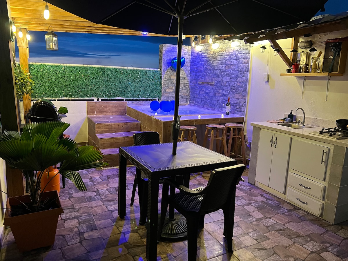 An outdoor space is presented with a covered dining area featuring a table and four chairs. A stone wall adds texture, while a hot tub is visible in the background alongside a grilling station. Soft lighting creates a relaxed environment for gatherings.