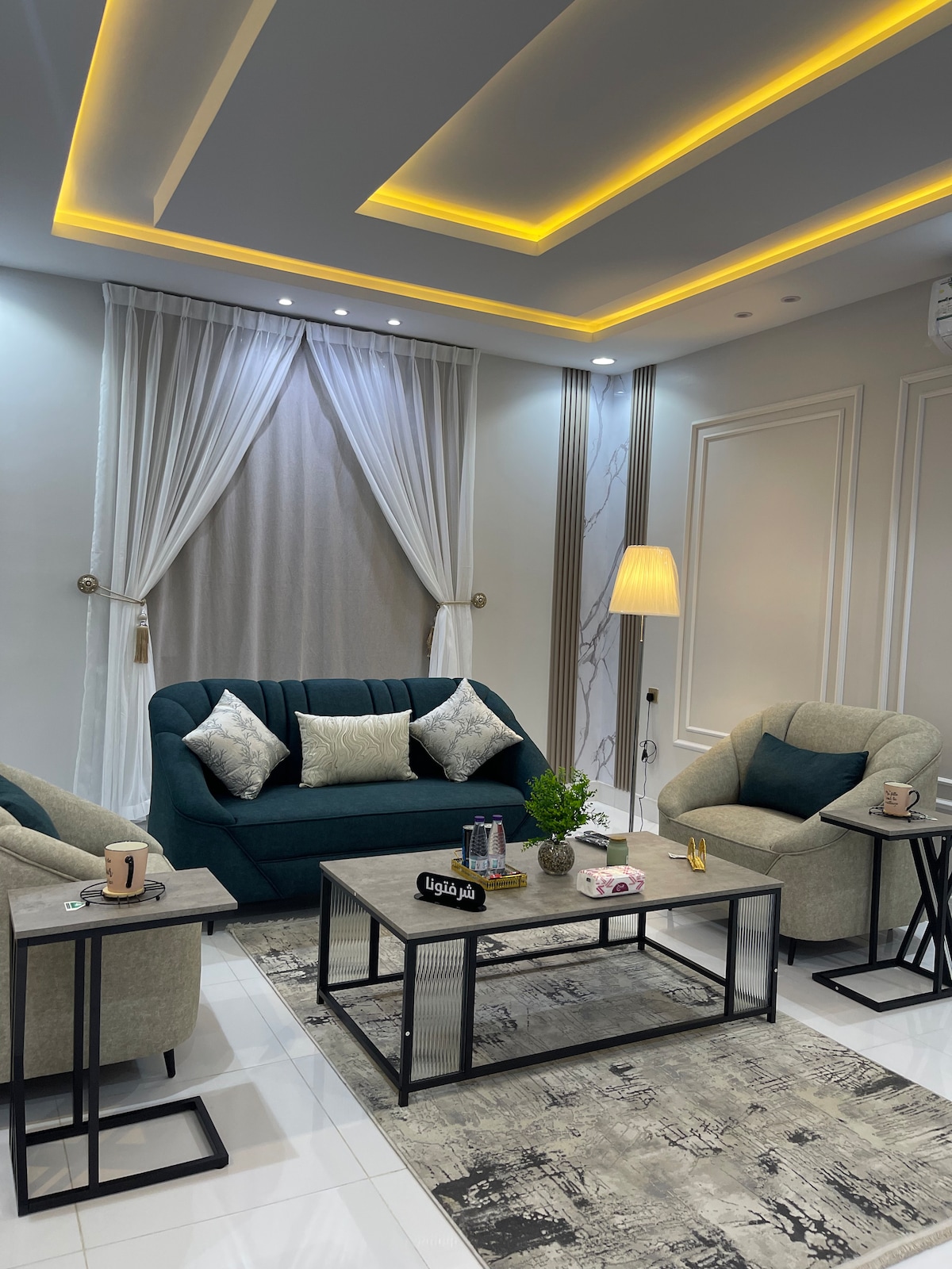 A modern living space features a comfortable sofa in deep green, complemented by two light gray armchairs. Two decorative side tables hold casual items. Soft lighting highlights the textured ceiling and floor, while sheer curtains frame a central window, adding elegance to the atmosphere.