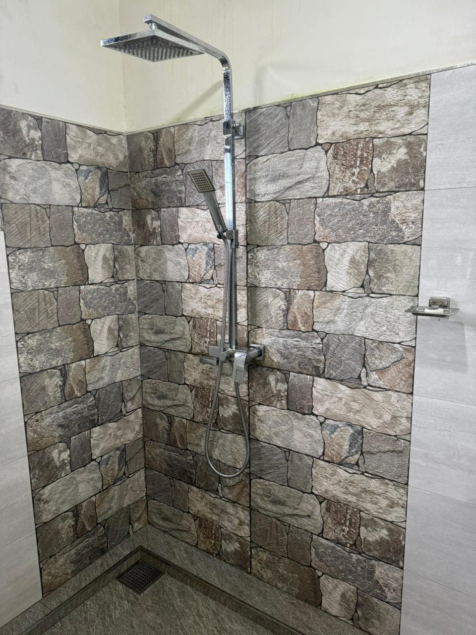 A modern shower area features a sleek showerhead mounted above a wall designed with textured stone tiles. A handheld shower attachment is positioned alongside. The floor is finished with a smooth, dark material, and a drain is visible in the corner.