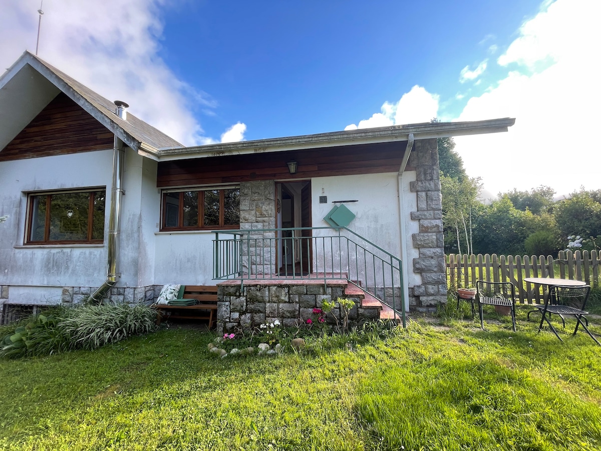 The entrance of the apartment is framed by a stone pathway leading to a wooden deck, with steps leading to the door. Lush green grass surrounds the area, complemented by a small garden with flowers. A dining set is positioned on one side.