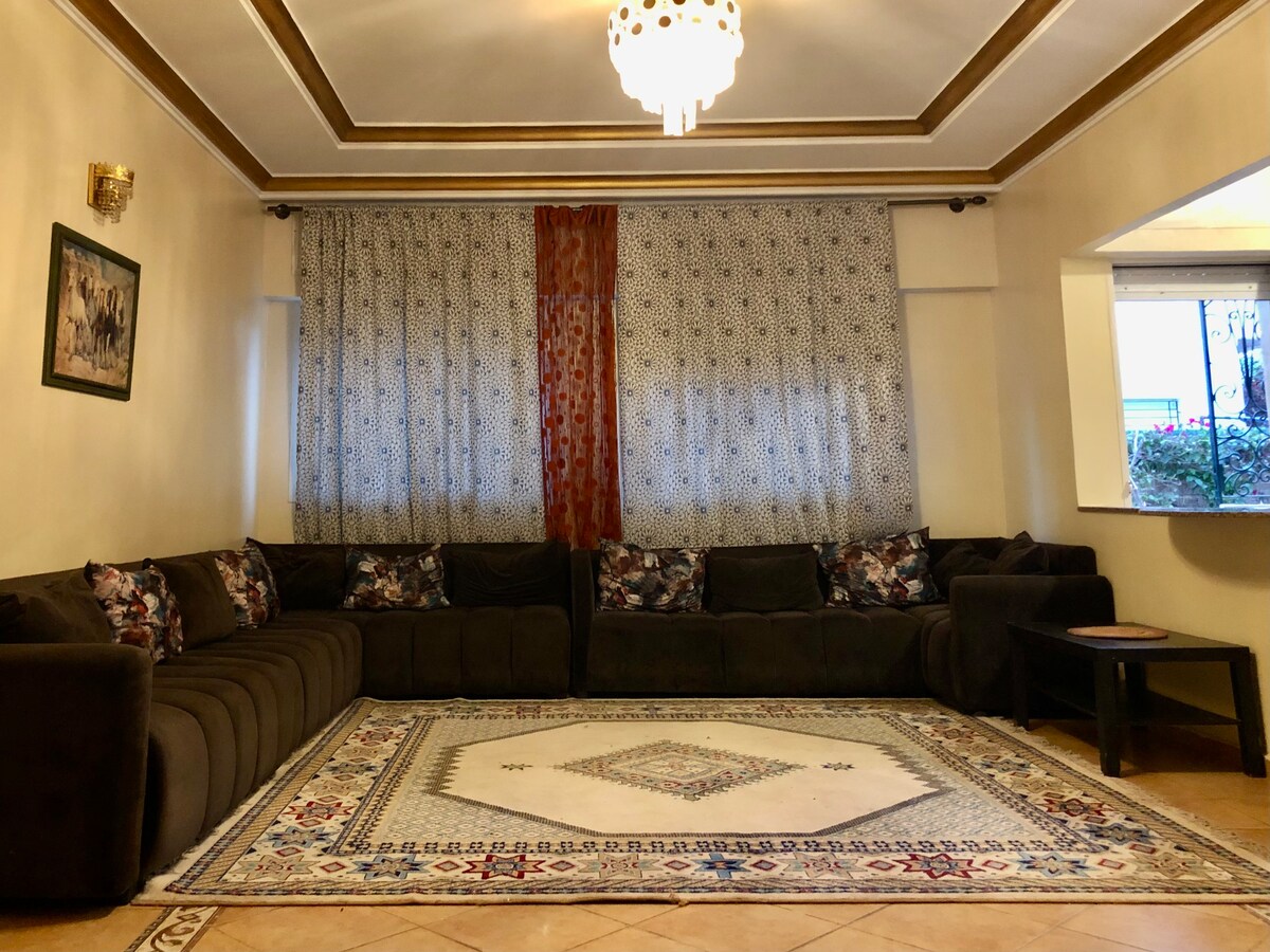 A large seating area features an L-shaped dark couch adorned with patterned throw pillows. A decorative area rug lies on the tiled floor, complementing the elegant design of the space. Natural light enters through a window partially covered by patterned curtains.