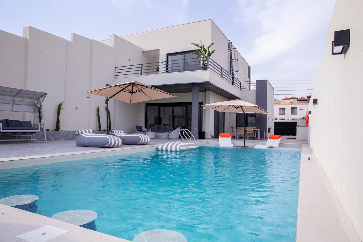 An expansive pool area features a large swimming pool with various seating options nearby. Lounge chairs and shaded areas provide comfort, while umbrellas offer protection from the sun. The modern villa's design is complemented by clean lines and a spacious patio overlooking the water.