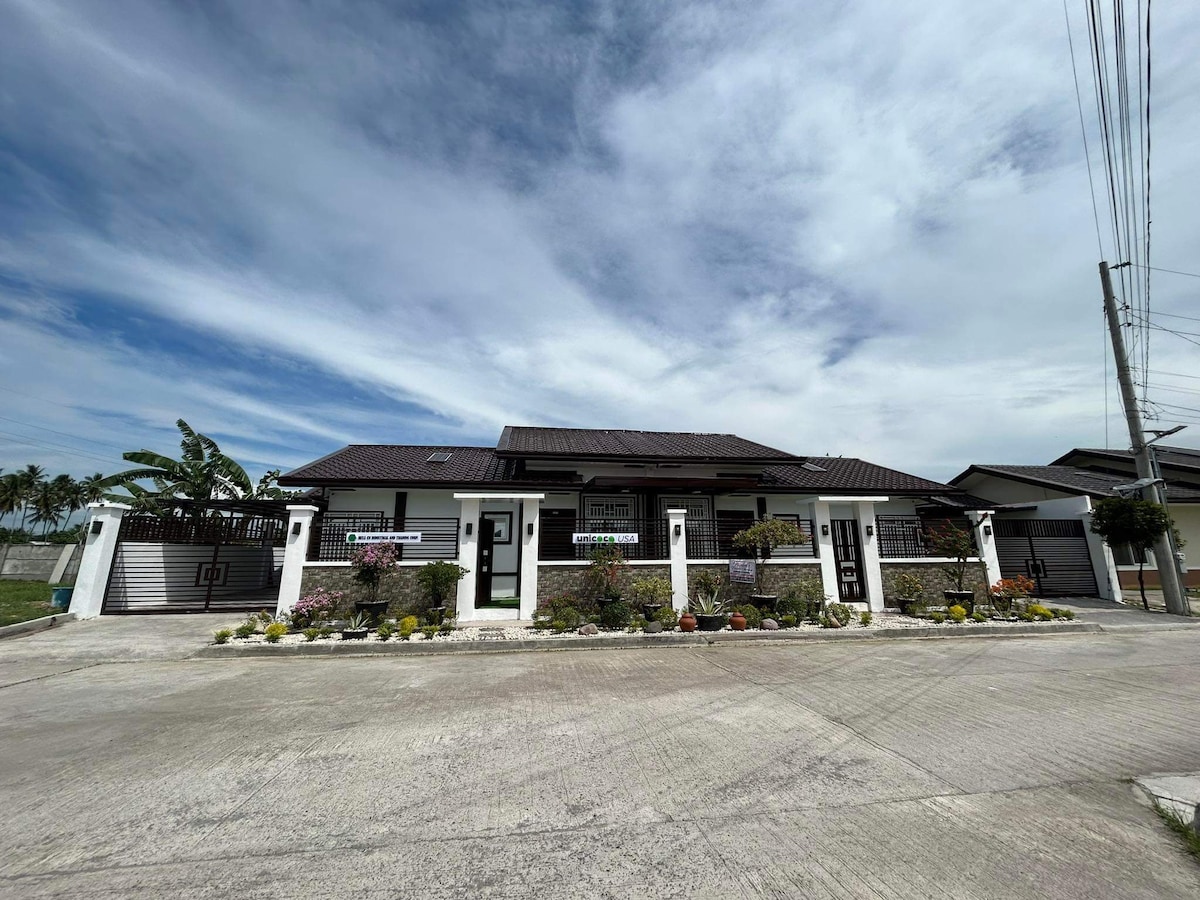 The exterior of the property showcases a distinct architectural design, complemented by a landscaped front yard with various plants and flowers. The building features multiple entrances and a tiled roof, while the surrounding area includes a paved driveway and nearby greenery.