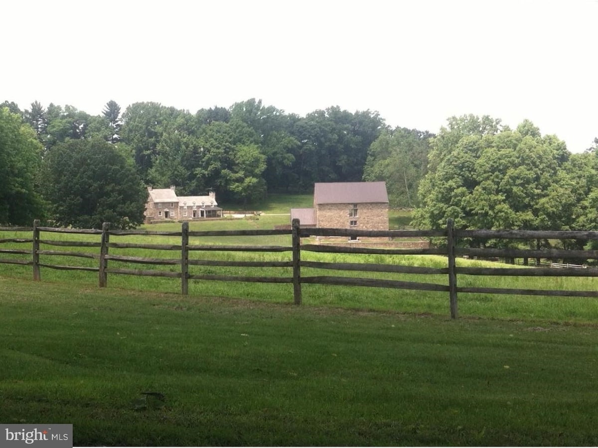 A historic stone building is surrounded by lush greenery, with a second structure visible in the background. A wooden fence lines the foreground, framing the expansive lawn and emphasizing the natural landscape that envelops the property.
