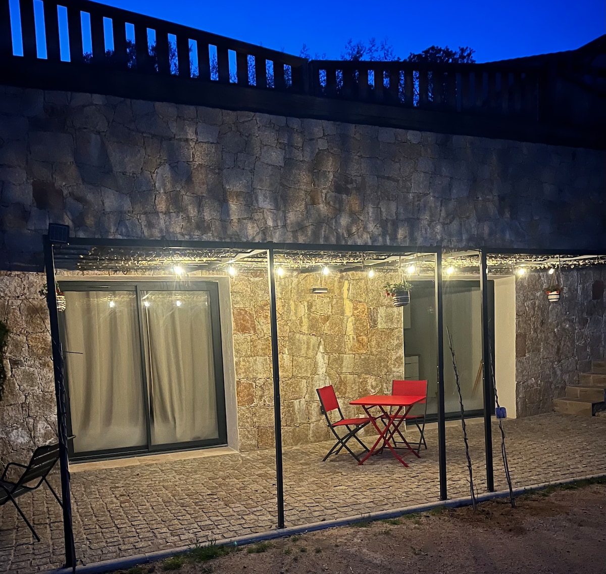 An outdoor patio is featured, illuminated by string lights. A small red table and matching chairs are set on a stone floor, surrounded by greenery. Large glass doors offer views into the interior space, contributing to a welcoming ambiance.