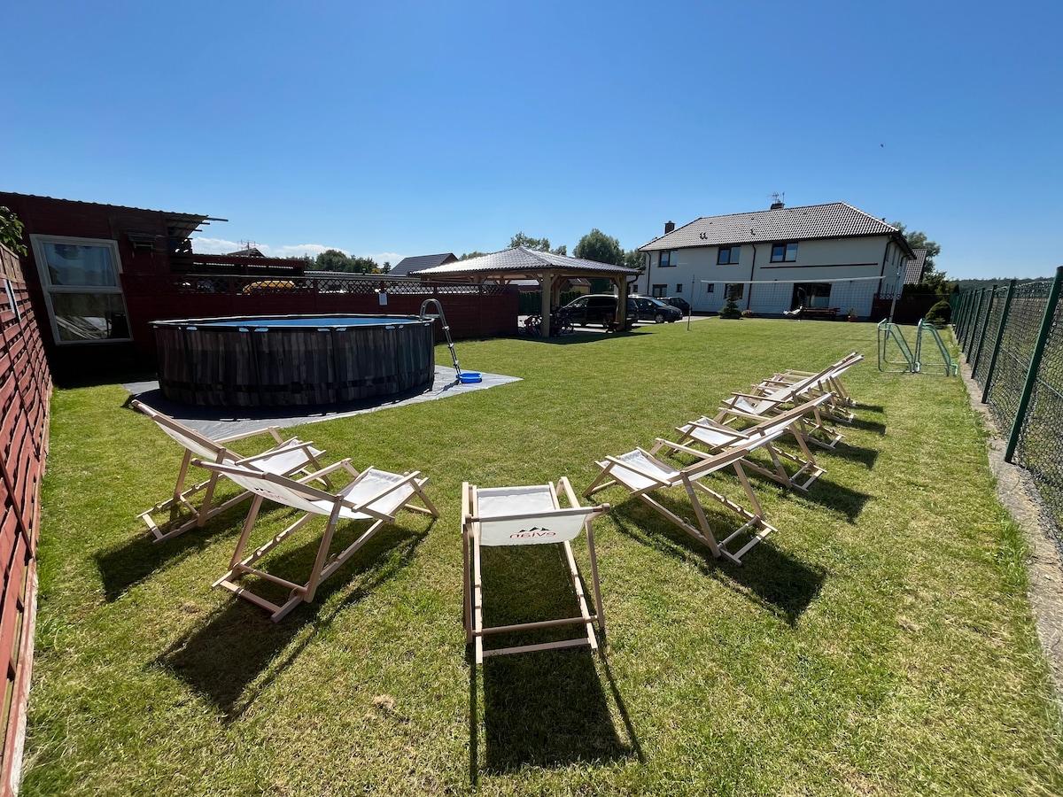 A spacious outdoor area is presented, featuring a well-maintained lawn surrounded by a fence. Several wooden deck chairs are arranged for relaxing in the sun. A wooden pool is visible, with a ladder leading into it, and a vehicle sits parked nearby.