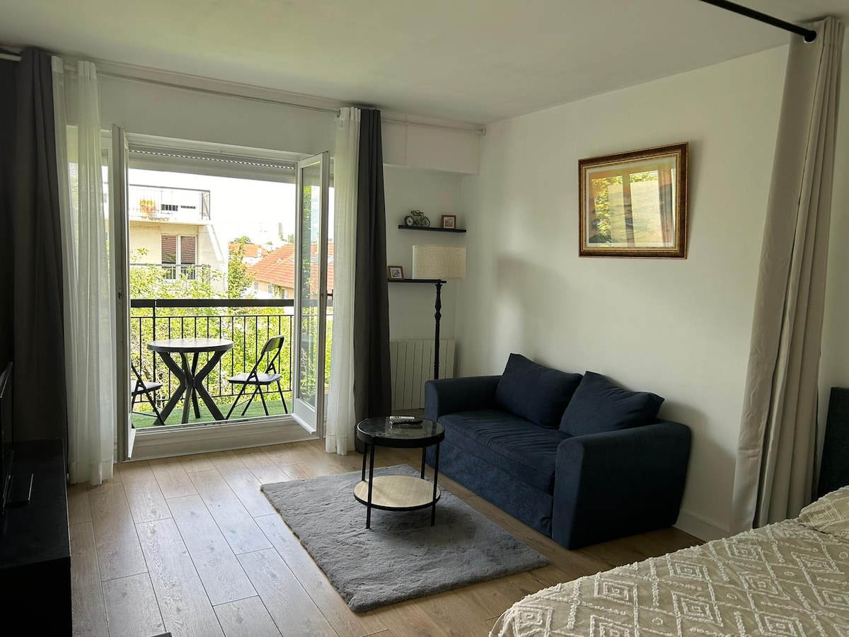 The living area features a comfortable dark blue sofa facing a flat-screen television. Natural light enters through large glass doors, leading to a small balcony with a bistro table and two chairs, overlooking a green garden area. A neutral rug lies under a round coffee table.