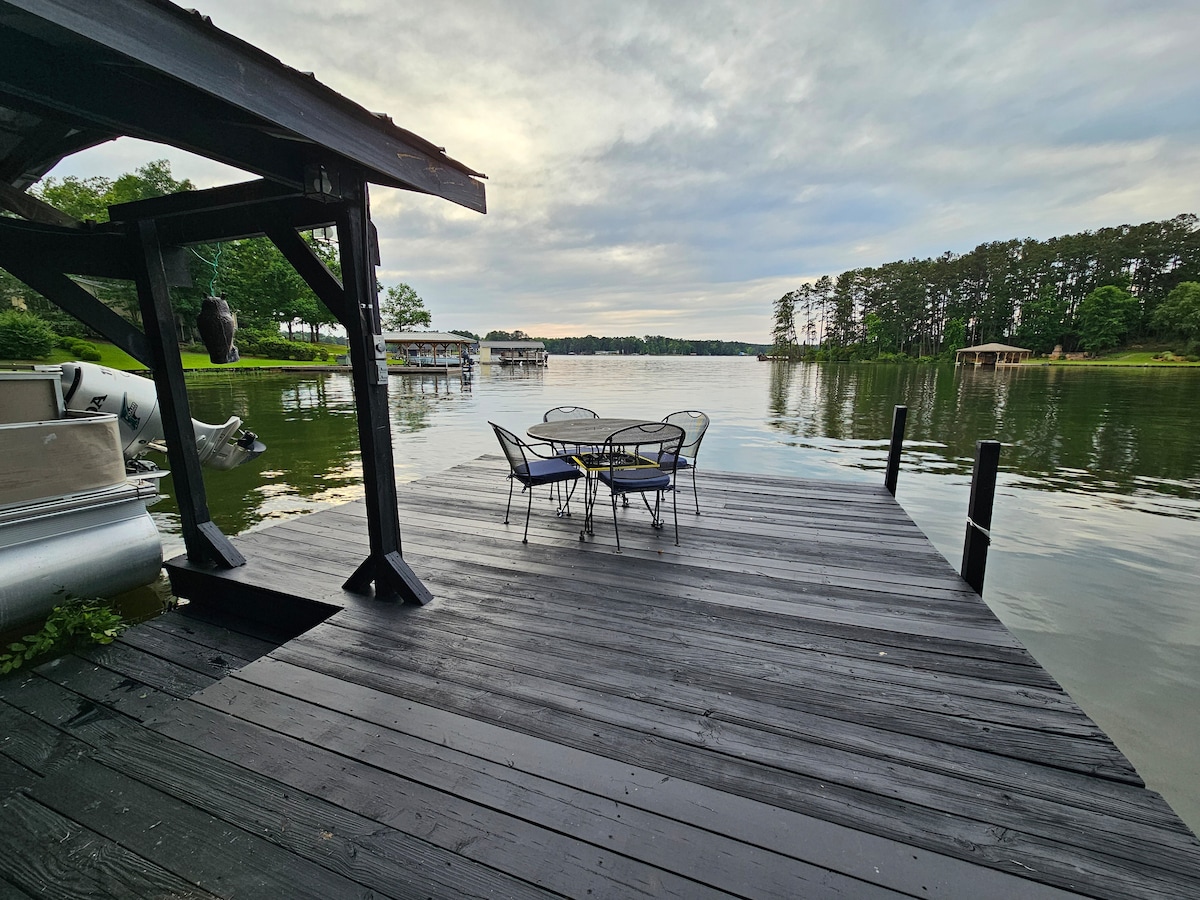 A wooden dock extends into Lake Sinclair, featuring a round table surrounded by four chairs. The calm water reflects the cloudy sky, with lush greenery lining the shore in the background. A boat is visible docked nearby, enhancing the lakeside atmosphere.