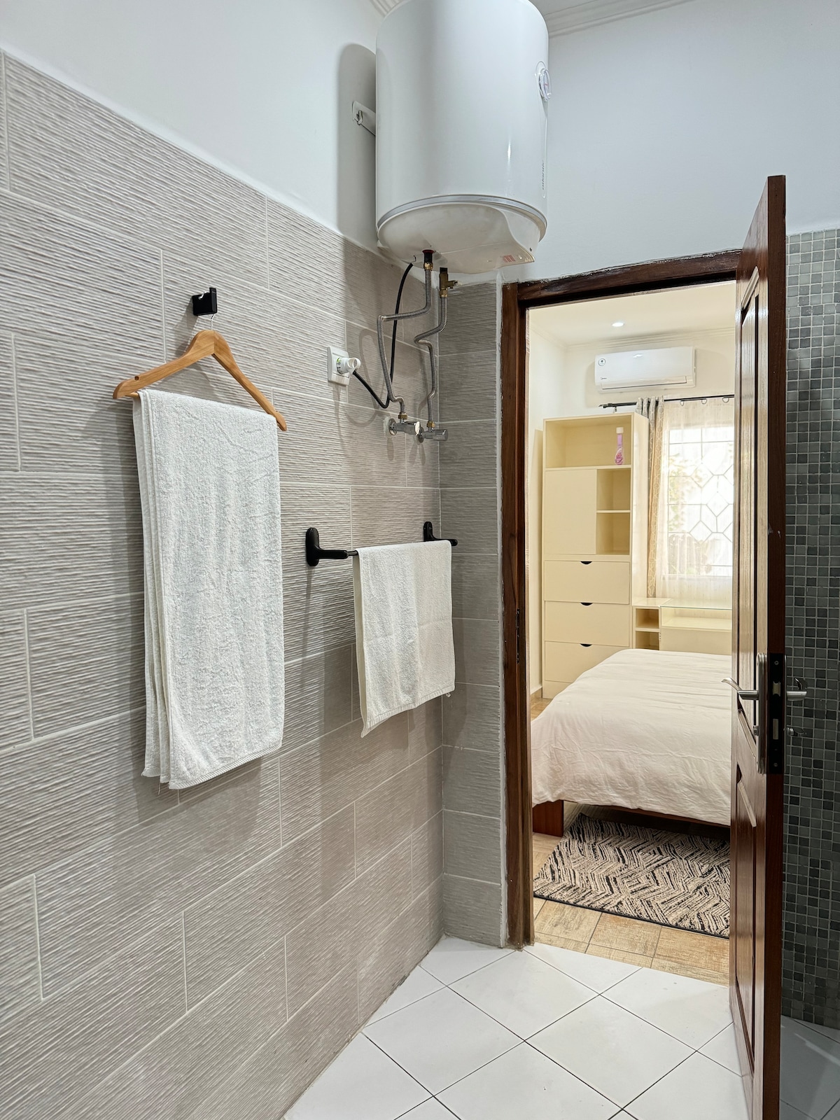 A compact bathroom area is showcased, featuring two neatly hung towels on black wall hooks. A water heater is installed above, and a doorway leads into a softly lit bedroom with light-colored furnishings, visible in the background.