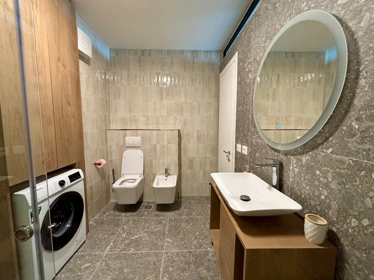 A modern bathroom is presented featuring a spacious shower area with glass walls, a large round mirror above a stylish sink, and wooden cabinetry. A washing machine is positioned beside a toilet and bidet, all set against elegant stone wall tiles.