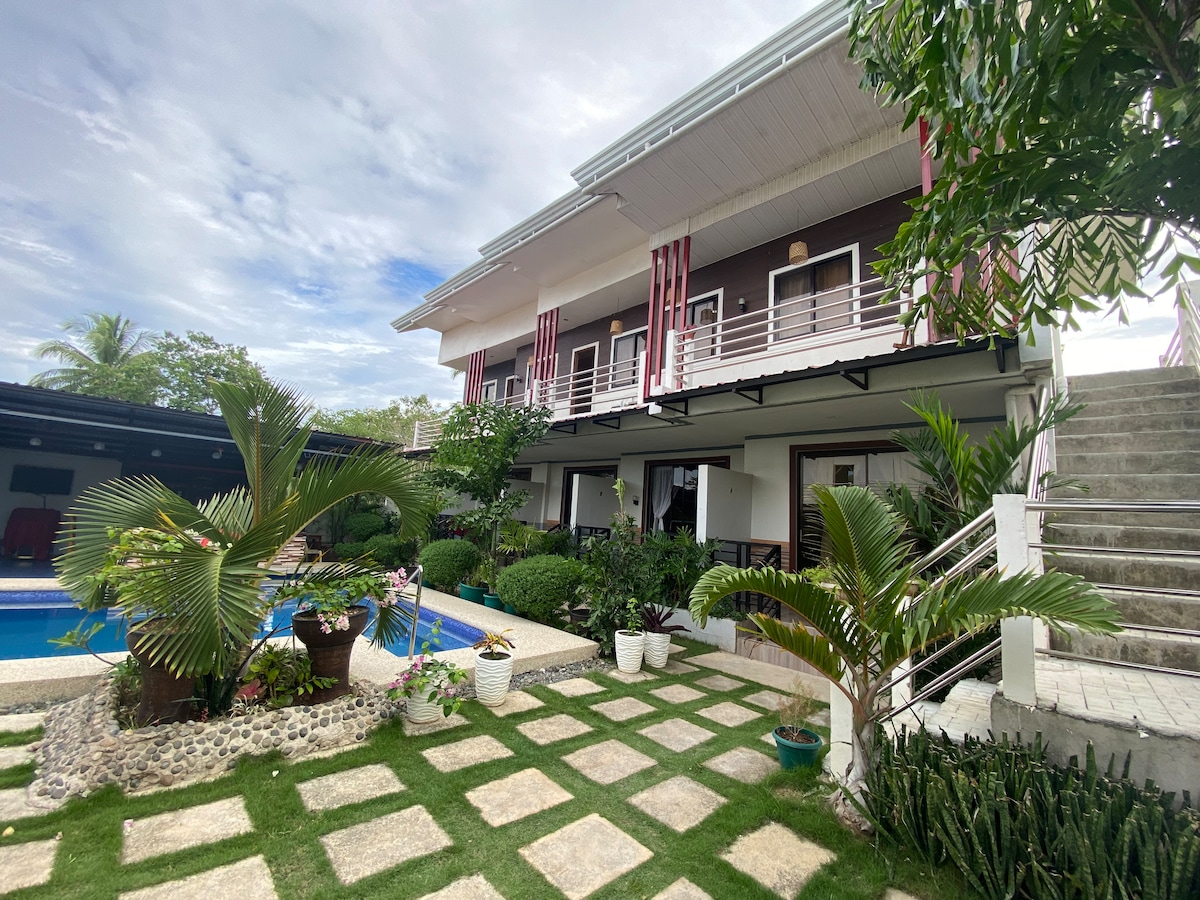 The exterior shows a two-story building with a balcony, framed by lush greenery and decorative plants. A well-maintained garden with stone pathways leads to a swimming pool, providing a serene outdoor setting.