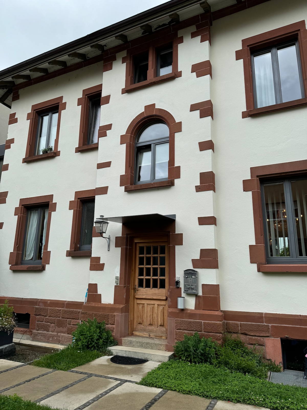 The exterior of a historic building is shown, featuring textured walls adorned with contrasting red and white stone. A wooden door is centrally located beneath an arched window. Windows are framed with dark trim, and a pathway leads to the entrance, complemented by small plants.