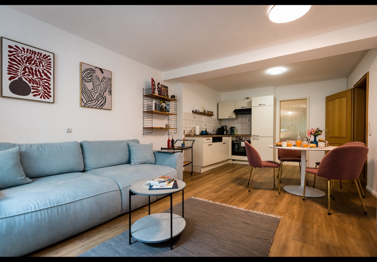 A cozy living area features a light blue sectional sofa complemented by a round coffee table. A dining space houses a small table with pink chairs, styled with a vase of flowers. The kitchen area is visible in the background, equipped with modern appliances.