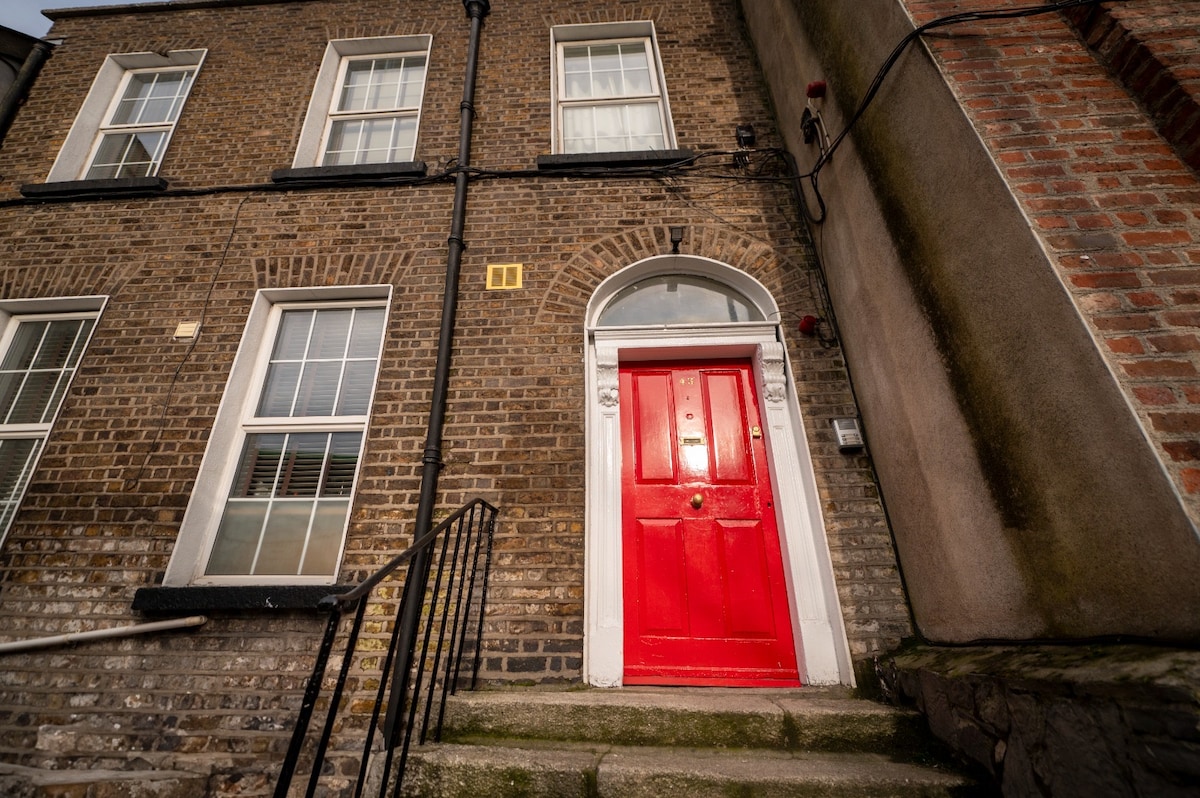 The exterior of the flat is highlighted by a vibrant red door at the entrance. The door is framed by elegant archways, with symmetrical windows on either side. Stone steps leading to the door are flanked by a handrail, providing a welcoming approach to the building.