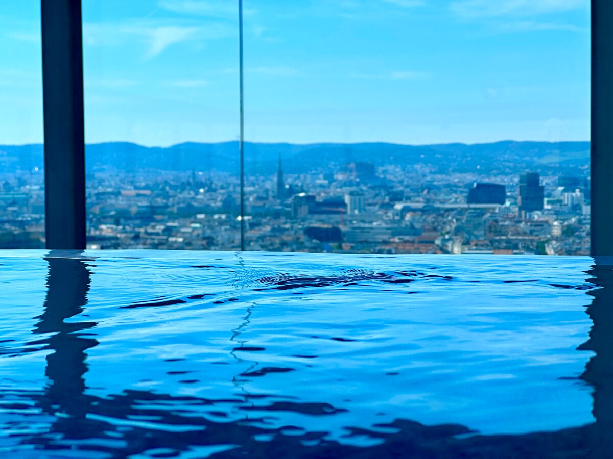 An infinity pool is visible, with clear, rippling water reflecting the sky. Expansive glass walls frame a panoramic view of the cityscape, revealing distant hills and buildings under a bright blue sky.