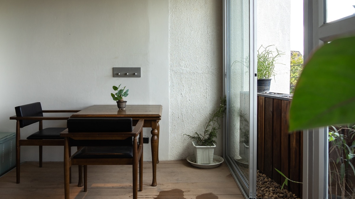 A wooden dining table is accompanied by two black chairs, positioned near a large glass window. Potted plants adorn the space, providing an element of greenery. Natural light illuminates the area, creating a serene spot for relaxation or dining.