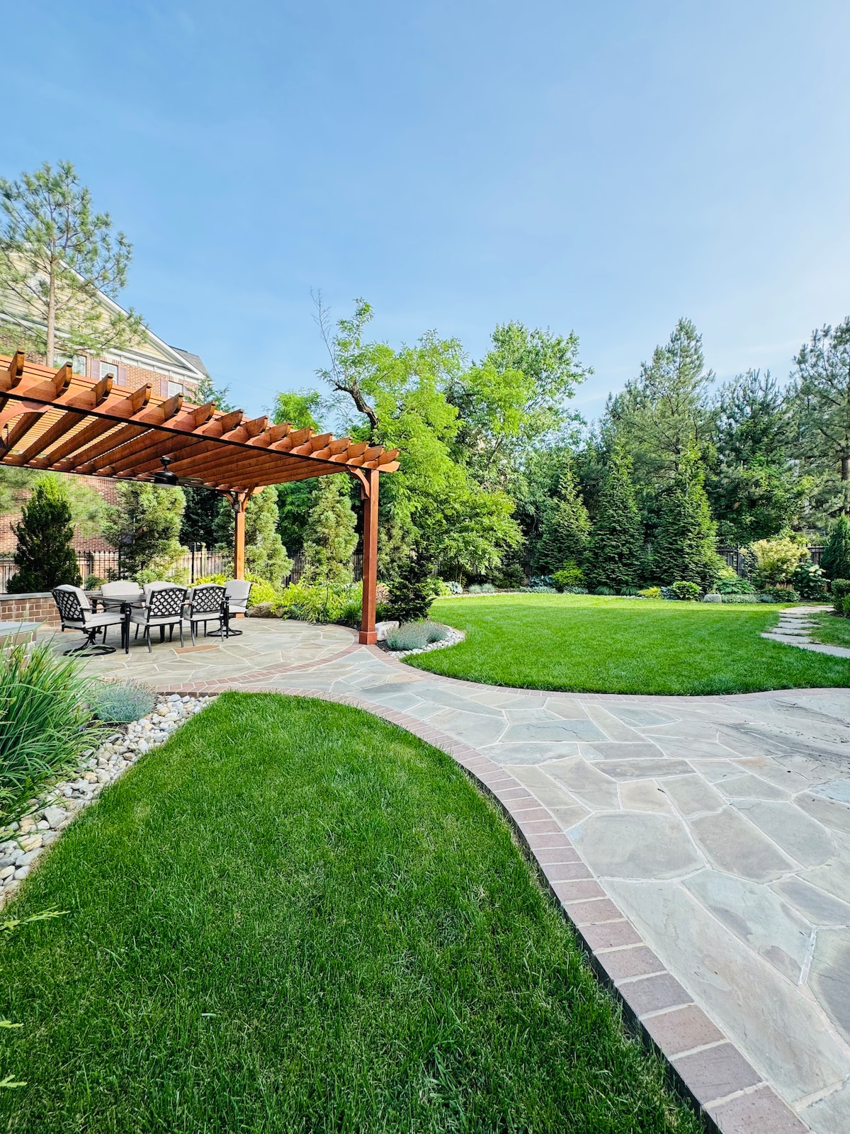 An outdoor space features a stone pathway winding through lush green grass, leading to a covered patio with a wooden pergola. Several outdoor chairs surround a table, nestled among trees and shrubs, creating a serene environment.