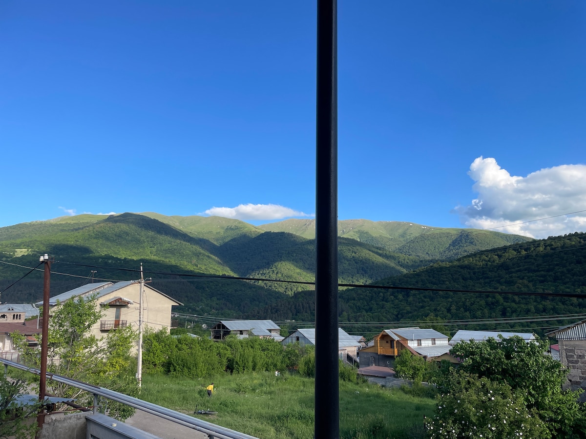 A scenic view of rolling mountains is framed by clear blue skies with fluffy white clouds. The foreground features various structures, including houses and greenery, creating a peaceful rural atmosphere.