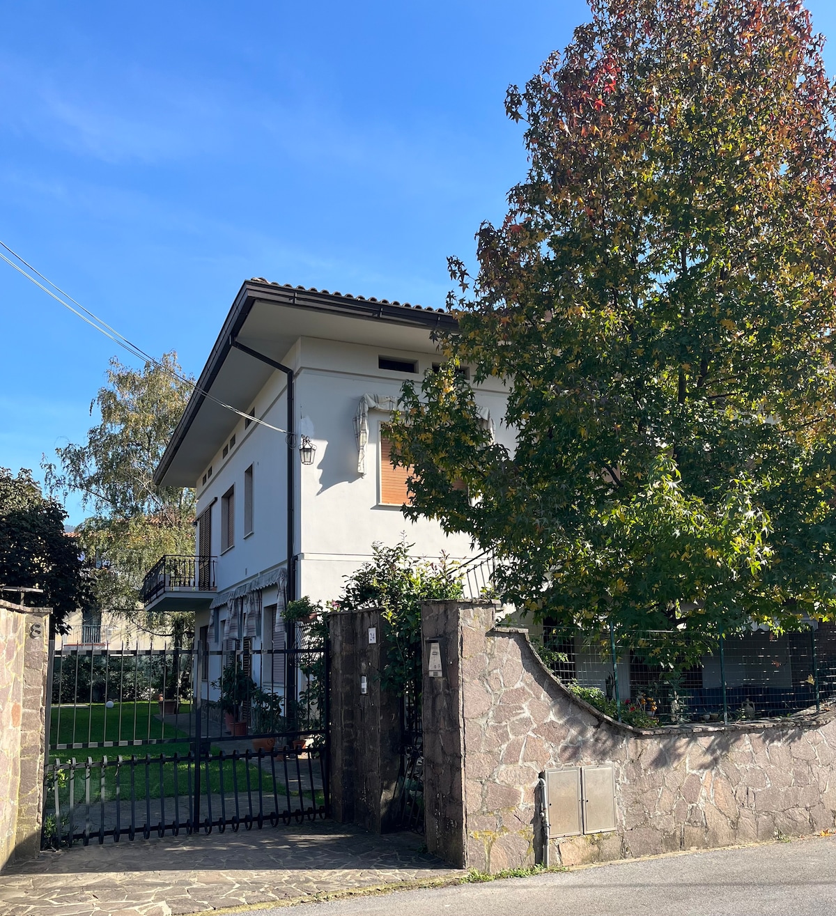 The exterior of the house showcases a two-story structure with a welcoming entrance. Lush greenery surrounds the property, including a large tree, creating a serene atmosphere. A gated pathway leads to the entrance, with clear blue skies visible above.
