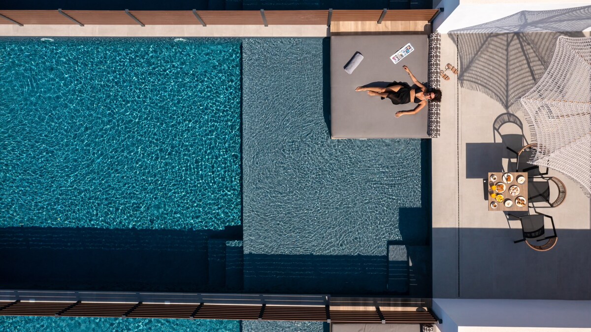 An aerial view reveals a private swimming pool surrounded by a sleek patio. A spacious lounging area is visible, along with a dining set arranged nearby. Warm sunlight reflects off the water, creating a serene and inviting atmosphere.