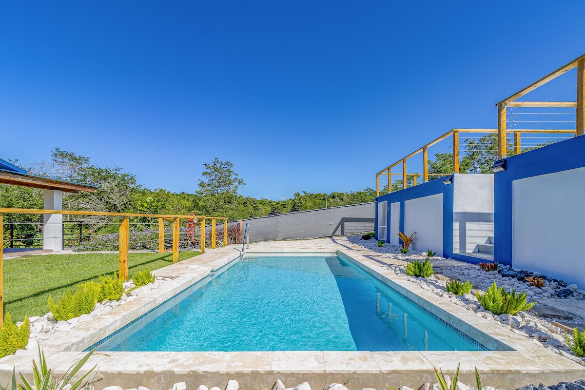 A clean, rectangular pool is positioned at the center of a sunlit outdoor area, surrounded by neatly arranged landscaping and local greenery. A wooden railing borders one side, while a view of the open sky adds a sense of spaciousness.