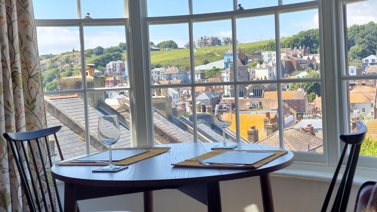 A round dining table is set beneath a large bay window, offering views of the picturesque Old Town rooftops and surrounding hills. Bright curtains with floral patterns frame the window, allowing natural light to fill the space.