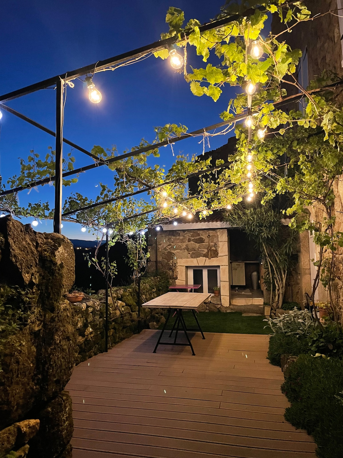 A charming terrace is illuminated by string lights, surrounded by lush vines. A wooden table is set on a wooden deck, providing an inviting outdoor dining space. The backdrop features stone walls and a built-in barbecue, enhancing the natural setting.