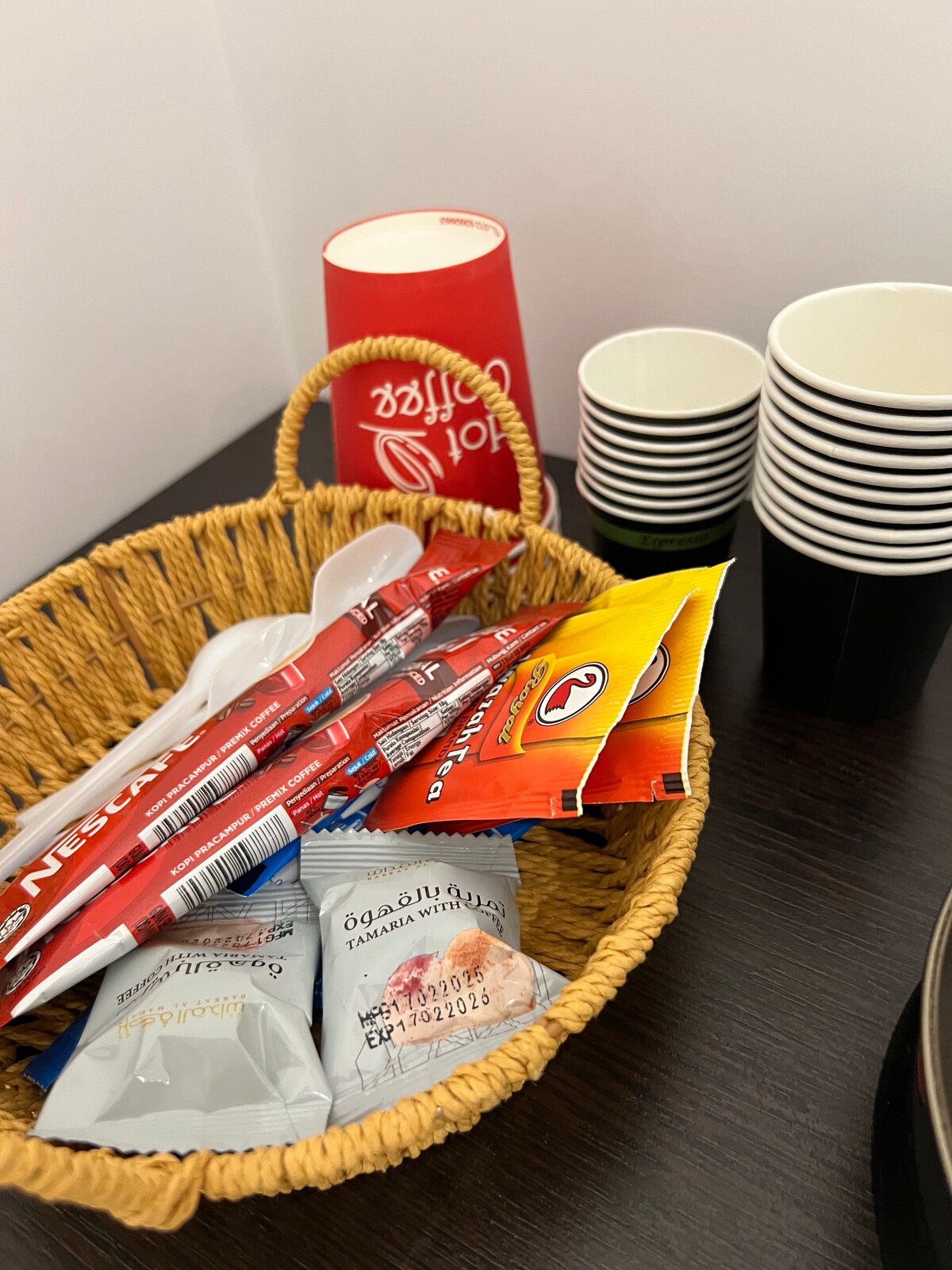 A wicker basket is filled with instant coffee packets and assorted snacks. Nearby, disposable cups are neatly stacked in black and red, creating a functional refreshment area. The dark wooden surface provides a contrasting backdrop, enhancing the organized presentation of items.