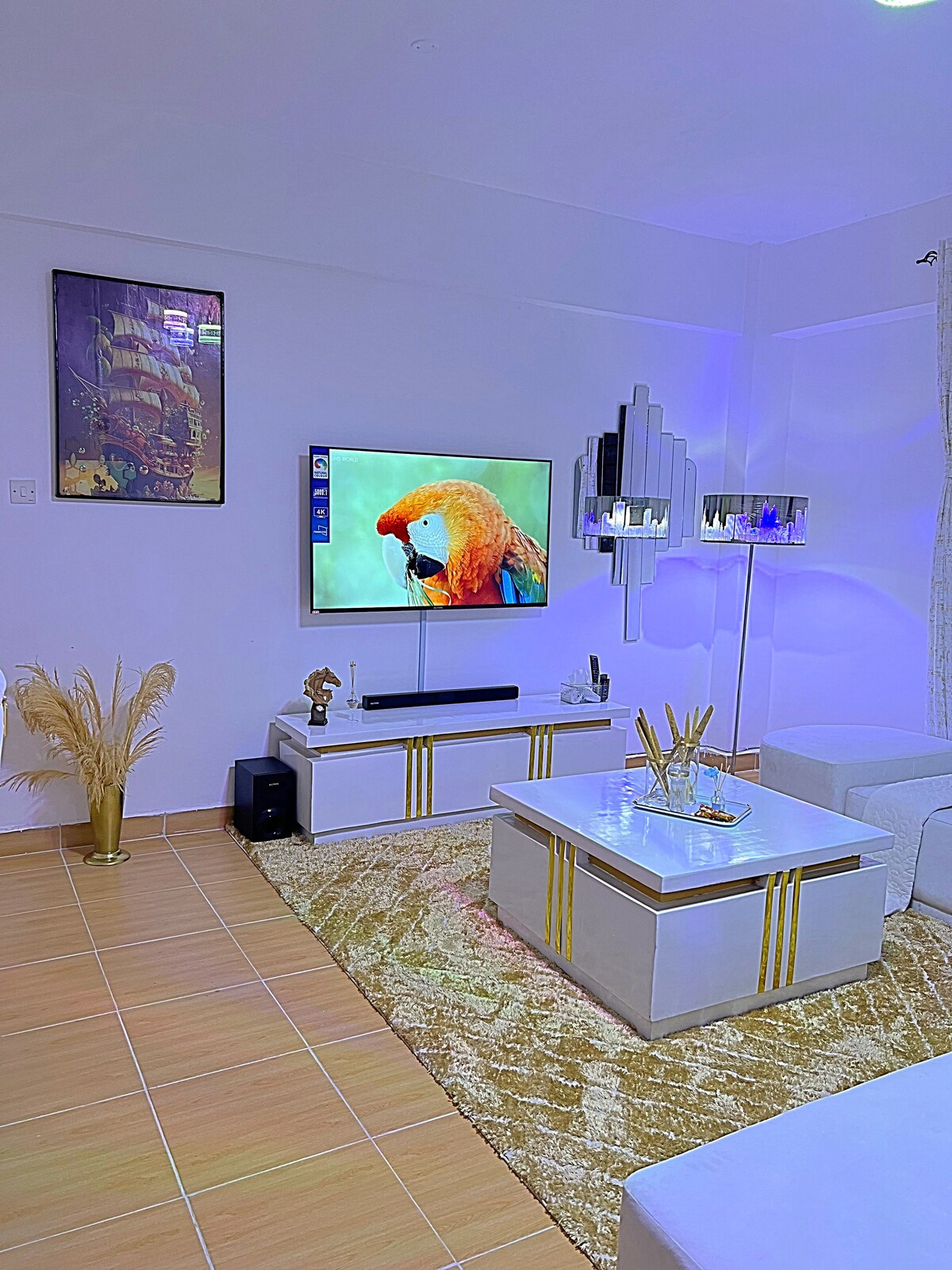 A spacious living area features a modern white entertainment unit with a large television mounted above it. A soft golden rug accents the floor, and decorative plants add a touch of vibrancy. Soft lighting creates a serene ambiance in the room.
