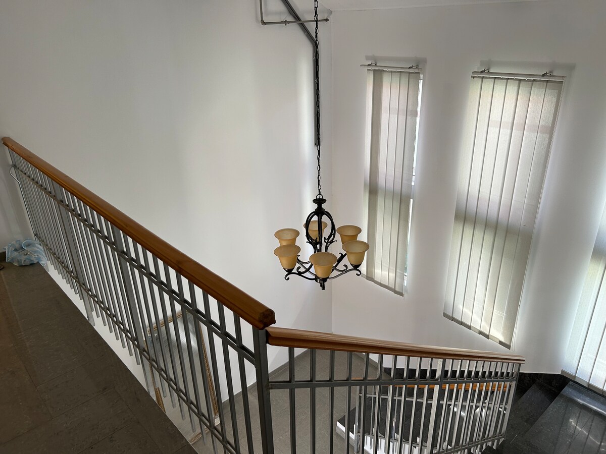 A modern staircase is shown, complemented by a stylish chandelier that hangs from the ceiling. Light-colored walls contrast with the dark staircase, while elongated window shades filter natural light from two vertical windows.