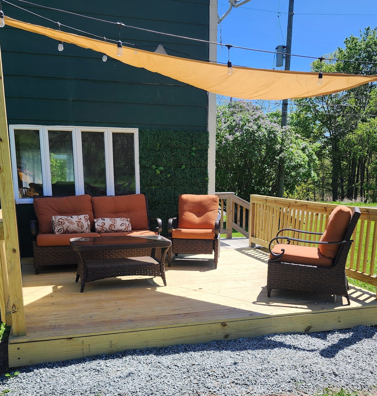 An outdoor deck is furnished with a seating area, including a comfortable couch and two armchairs, all with orange cushions. A glass-top coffee table is positioned at the center. A shade sail provides coverage, while greenery and gravel can be seen in the surrounding area.