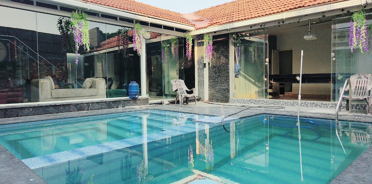 A private pool glistens under sunlight, surrounded by modern architecture. Large glass doors provide views into the spacious interiors. Lush greenery and hanging flowers add subtle color, while comfortable seating is positioned nearby, creating an inviting space for relaxation and leisure.