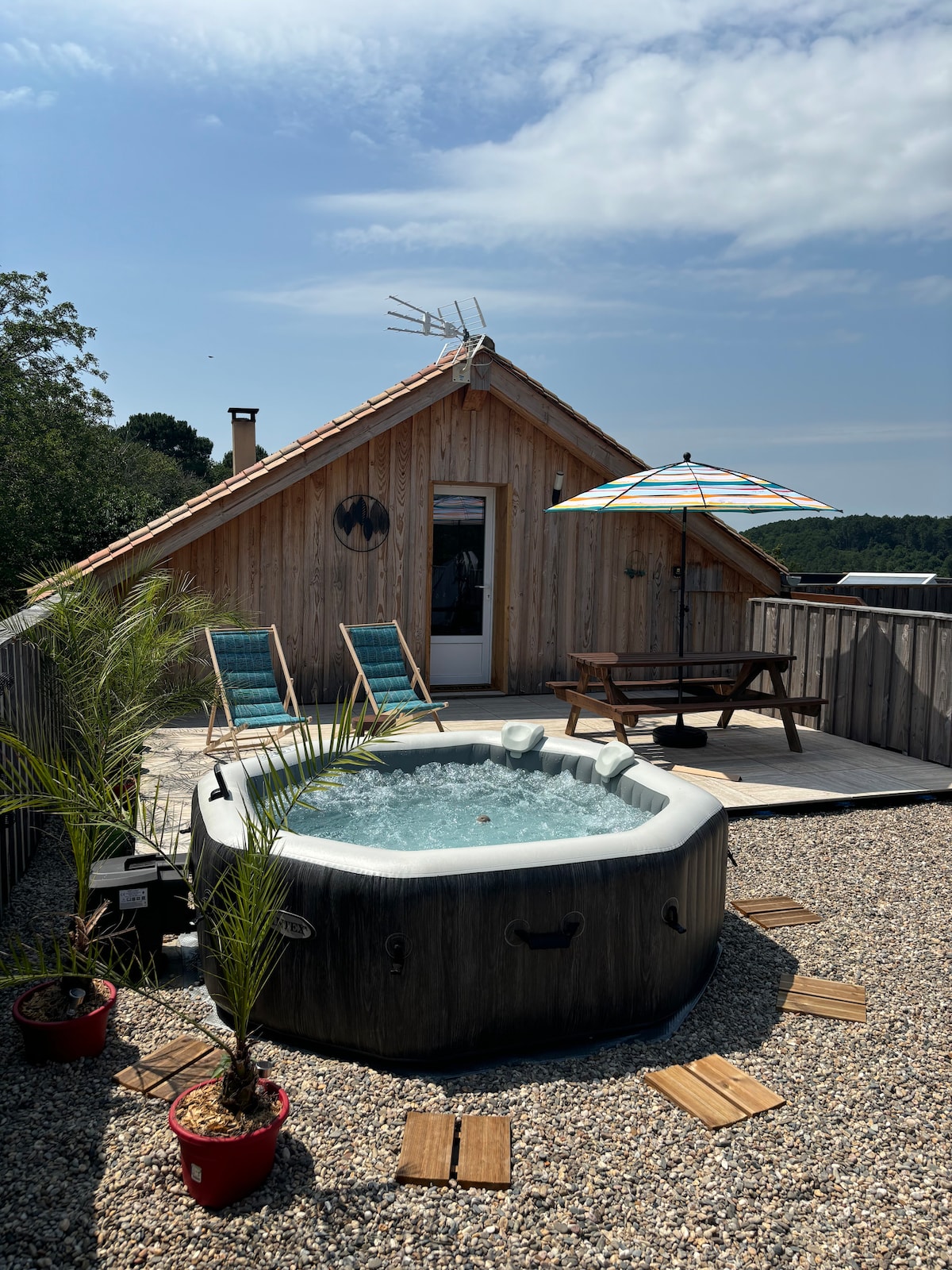 A private spa surrounded by comfortable lounge chairs and a shaded patio umbrella is featured on the rooftop terrace. Potted plants are placed around the area, with a picnic table present for outdoor dining. The wooden structure and natural surroundings create a serene environment.