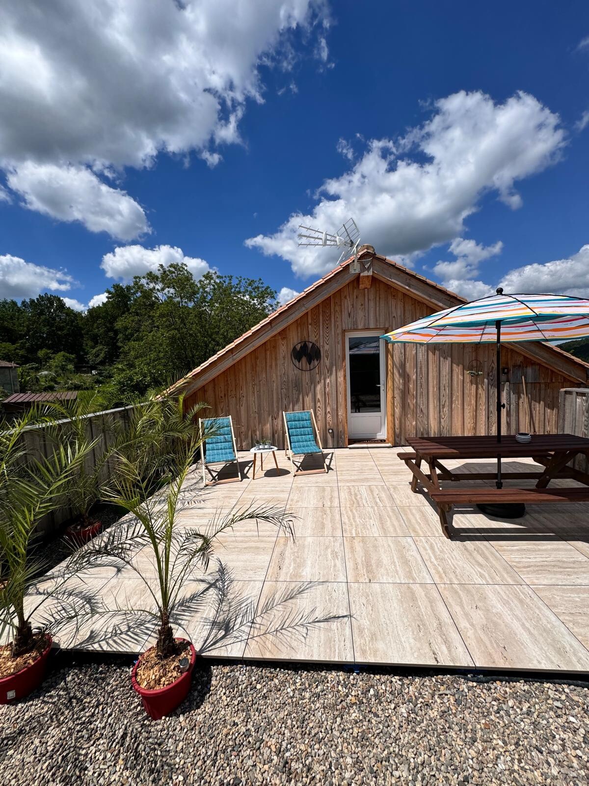 An outdoor terrace features a spacious, tiled area with two lounge chairs and a large umbrella providing shade. Potted palm plants add a touch of greenery, while a wooden table and bench invite relaxation. The building’s wooden exterior complements the natural surroundings under a bright blue sky.